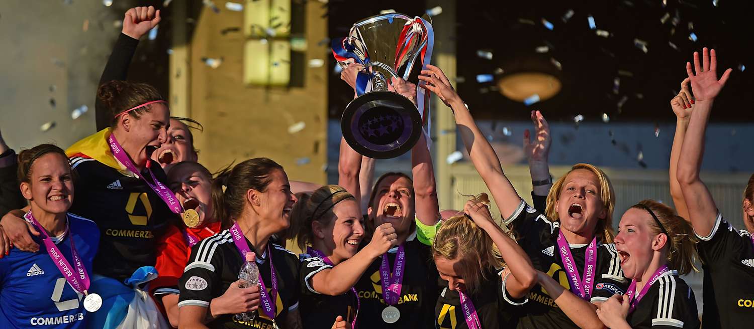 Spielerinnen der Eintracht Frankfurt Frauenmannschaft jubeln in dunklen Trikots über ihren Triumph in einem europäischen Wettbewerb. In der Mitte des Bildes wird der Pokal mit silbernem Glanz, verziert mit blauen, weißen und roten Bändern, in die Höhe gestreckt. 