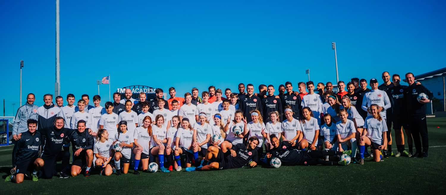 Vielen Menschen stehen gemeinsam und posieren für ein Gruppenbild. Darunter ehemalige Spieler von Eintracht Frankfurt wie Filip Kostic, Legenden des Vereins wie Karl-Heinz Körbel und Kinder eines Fußballschulencamps in den USA.