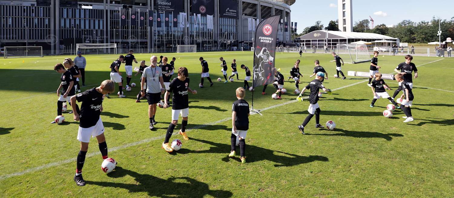 Viele Kinder machen sich gemeinsam im Mittelkreis warm. Jedes Kind hat einen Ball am Fuß, am Mittelkreis befindet sich eine Fahne der Eintracht Frankfurt Fußballschule. Trainer Alexander Conrad befindet sich ohne Ball im Mittelkreis und schaut sich die Kids aus nächster Nähe an. Die Einheit findet am Trainingsplatz am Fuß des Deutsche Bank Park statt.