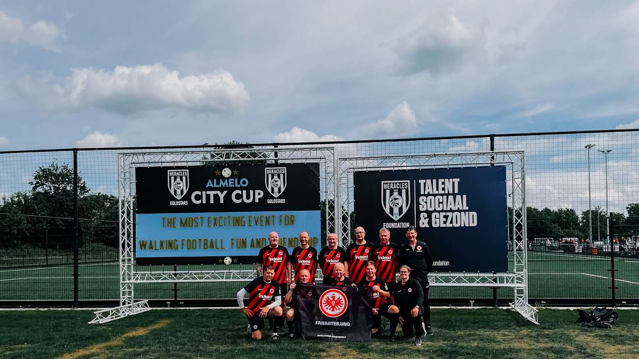 Die Walking Football Mannschaft der Eintracht macht mit Trainerin Steffi ein Gruppenbild vor einem Zaun mit zwei großen Bannern.