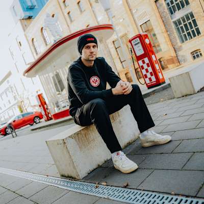 Sitzend, schräg von links: Mann mit schwarzer Eintracht Frankfurt Mütze und schwarzem Pullover mit SGE-Logo sitzt auf einem Stein. Fisherman Beanie 1899