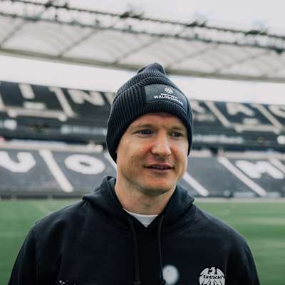 Mann mit schwarzer SGE-Beanie mit Waldstadion-Logo, schwarzem Eintracht Hoodie vor Stadiontribüne. Beanie Waldstadion Basic