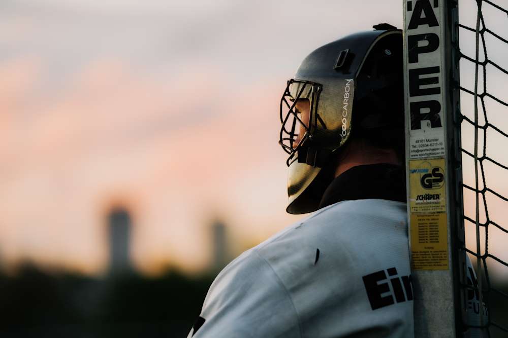 Torwart der Hockey-Herren schaut in Richtung Hockeyplatz. Er trägt ein weißes Trikot und einen typischen Feldhockey-Helm.